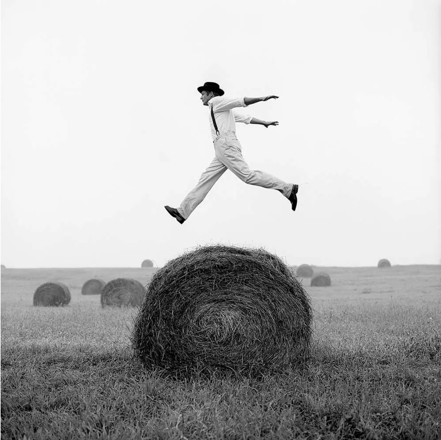 A man in a bowler hat jumping over a hay bale